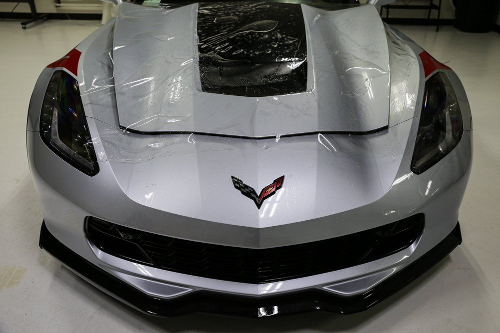Silver Corvette front end with protective wrap, black stripes, red accents, and black front splitter.