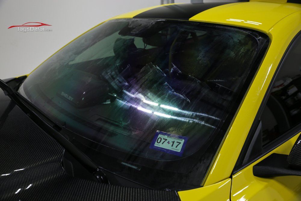 Damaged windshield on a yellow sports car. Black hood with racing stripes, windshield has multiple cracks.