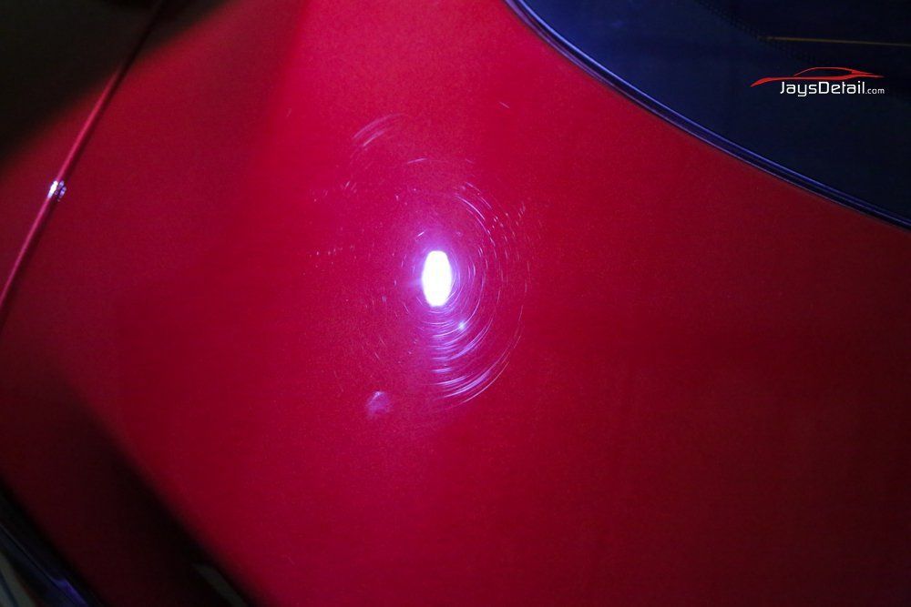 Red car paint with circular scratches reflecting light.