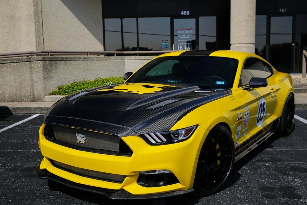 Yellow and black Ford Mustang parked in front of a building with black rims and a carbon fiber hood.