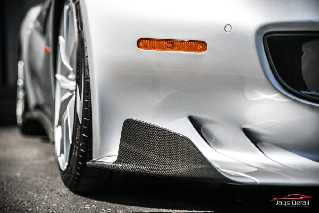 Silver car front with carbon fiber splitter, orange reflector, and alloy wheel.