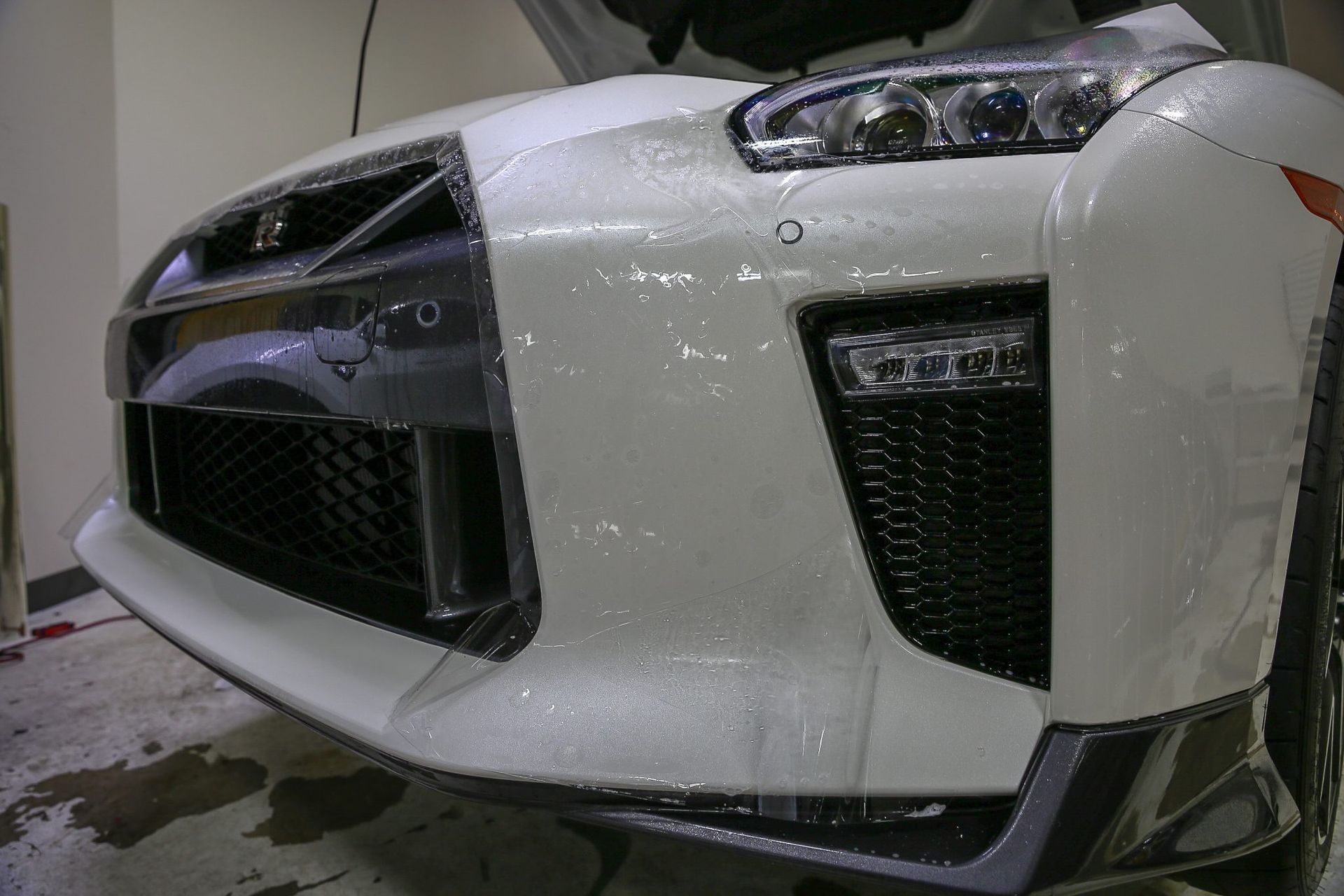 White car front bumper with protective film applied. Black grilles and headlights visible.