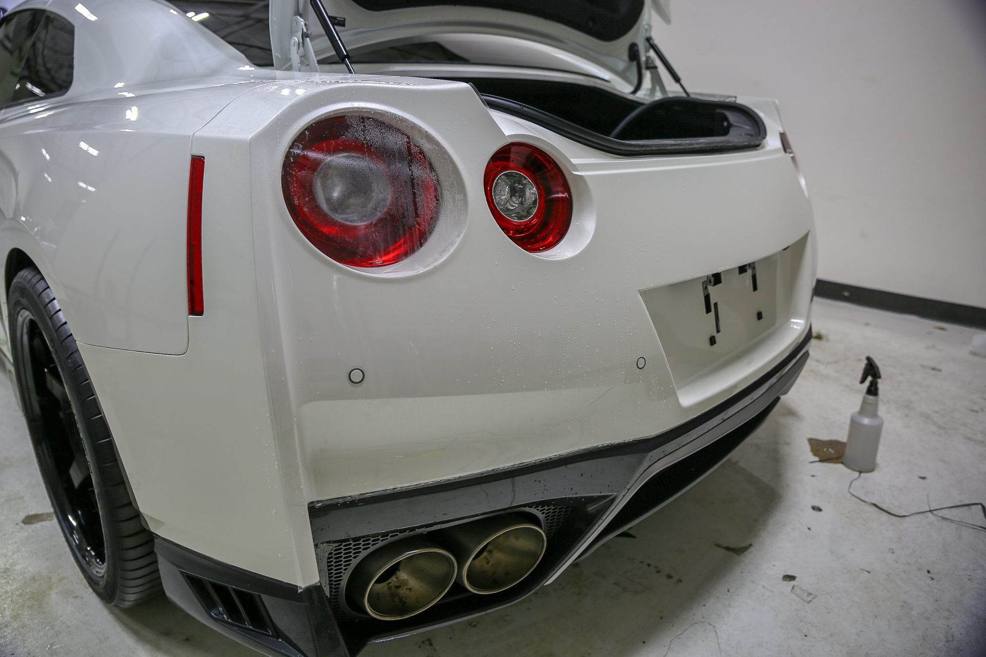 Rear view of a white Nissan GT-R with open trunk, black wheels, red taillights, and dual exhaust.