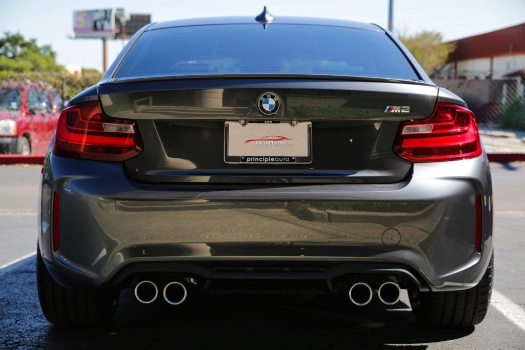 Rear view of a gray BMW M2 with dual exhaust pipes, parked outdoors in sunlight.