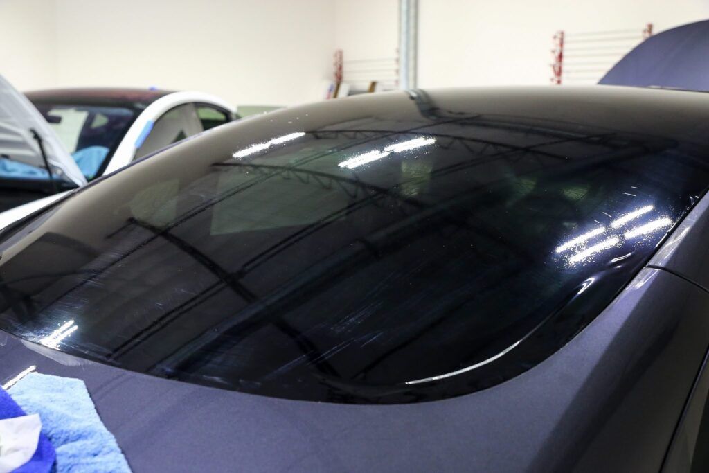 Dark-tinted car windshield reflecting the interior of a garage, partially covering a blue car.