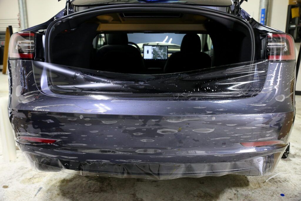Dark gray Tesla with the rear trunk open, covered in protective film indoors.