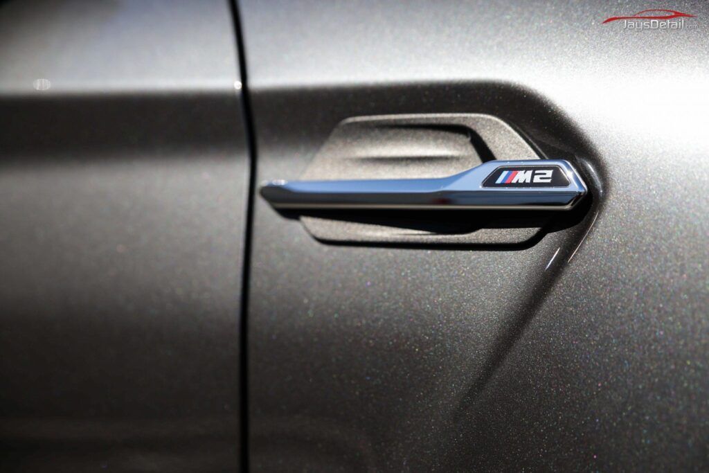 Close-up of a grey BMW M2 door handle with chrome accents and M2 logo.
