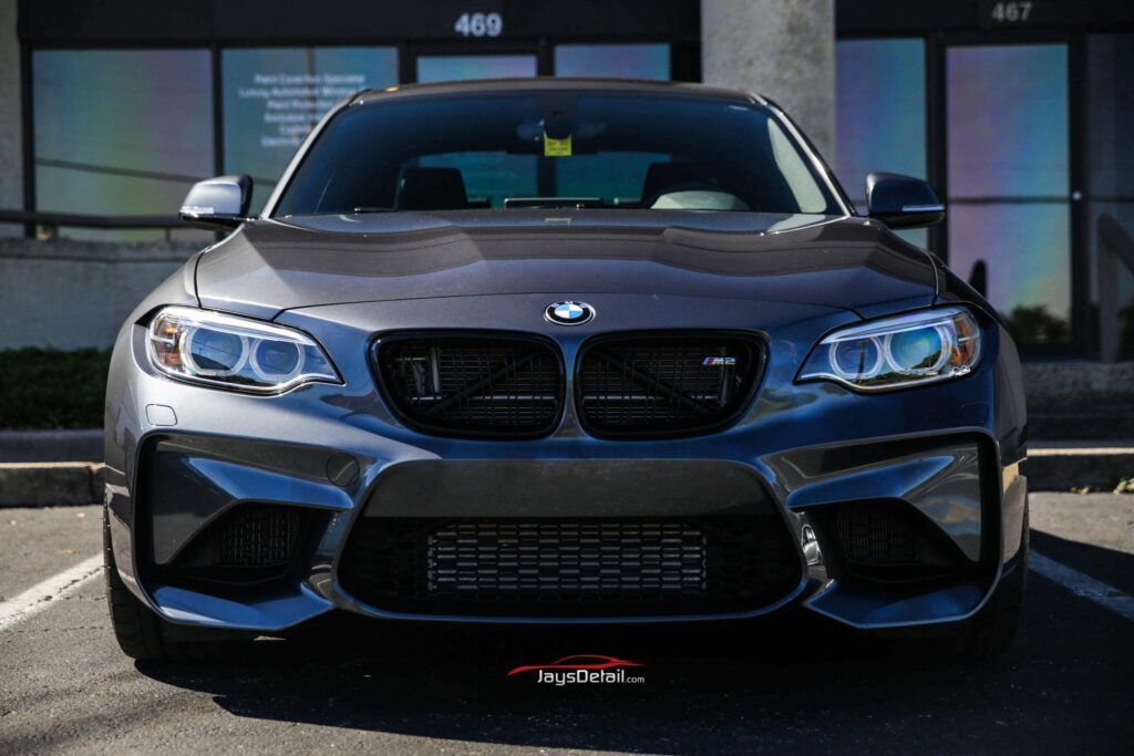 Dark gray BMW M2 coupe parked in front of a building with black grilles and blue-tinted headlights.