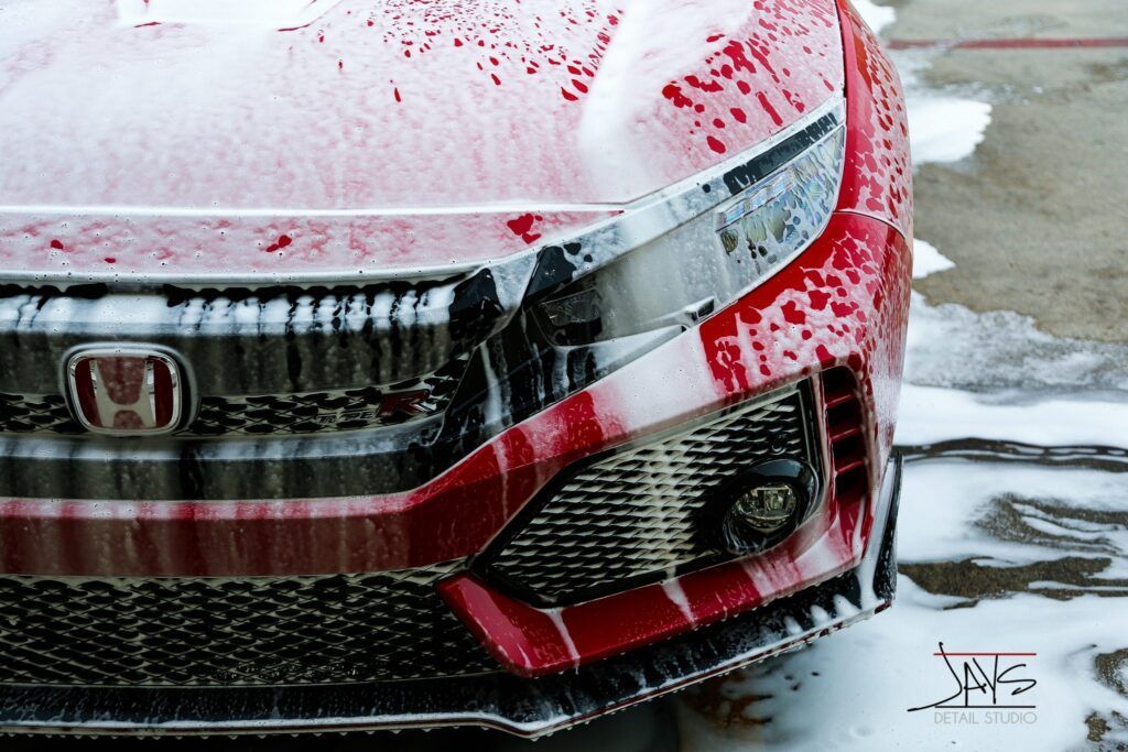 Red Honda car being washed with soap, covered in white foam.