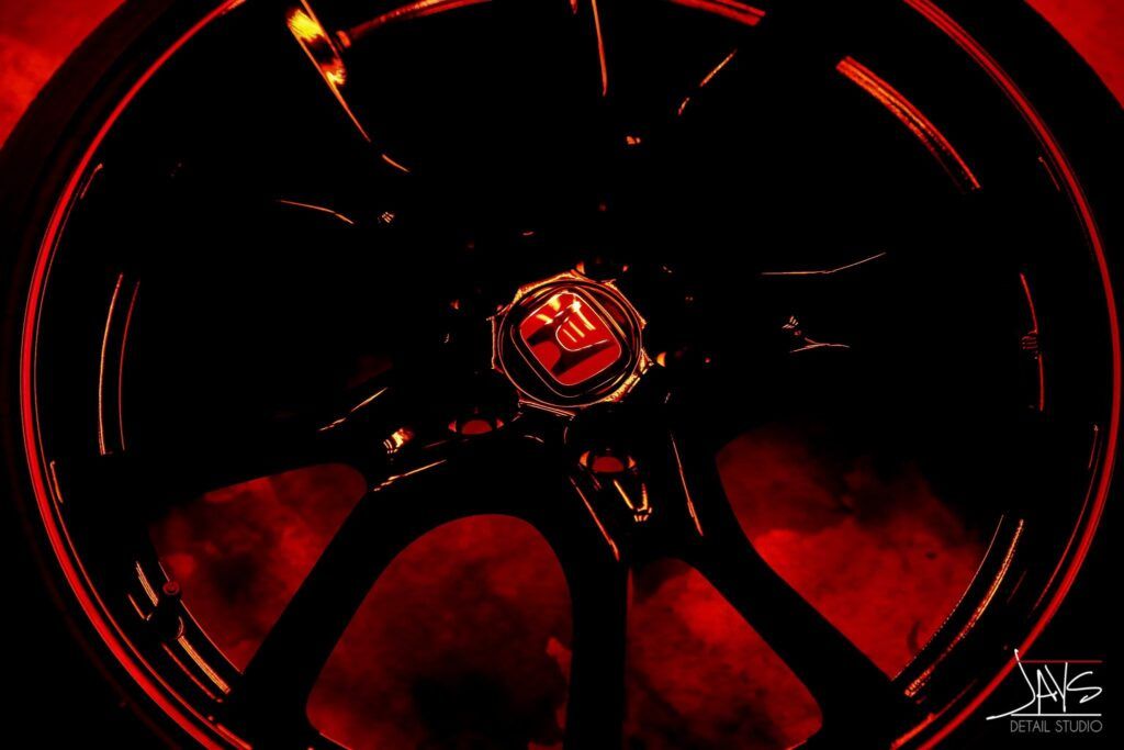 Black car wheel rim against a dark red background, the center features a logo.