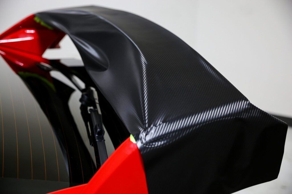 Carbon fiber rear car spoiler, black, over a red car body, near a window.