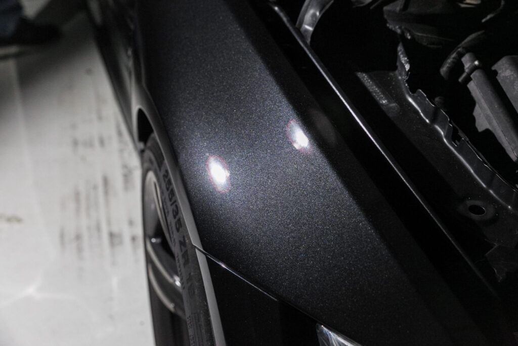 Dark gray car fender with shiny highlights near the open hood in a bright setting.