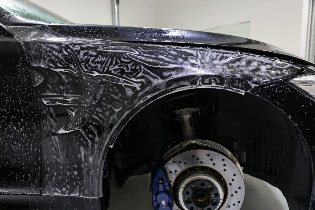 Black car wheel well being washed with soapy foam; showing brake disc and blue caliper.