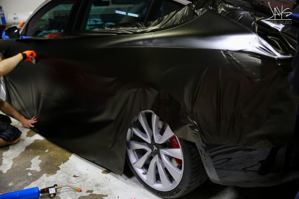 A person wrapping a black car with black vinyl, near the rear wheel.