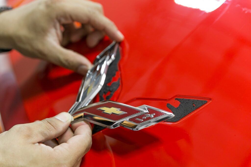 Hands placing a silver and red Corvette emblem on a red car panel.