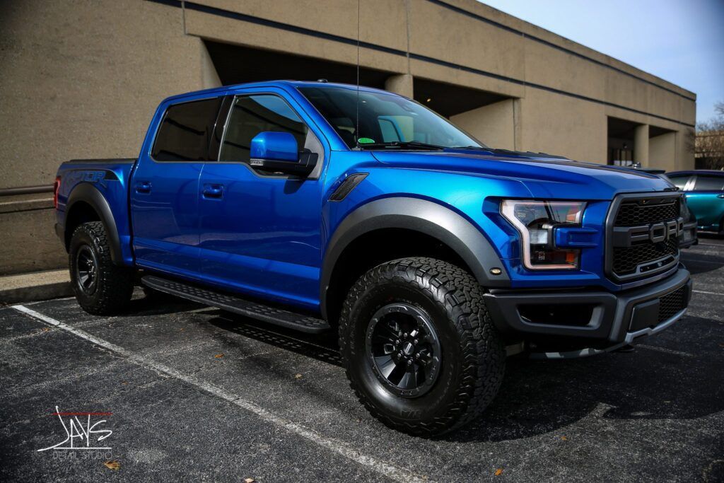 Blue Ford Raptor pickup truck parked in a lot, black wheels and trim, near a beige building.
