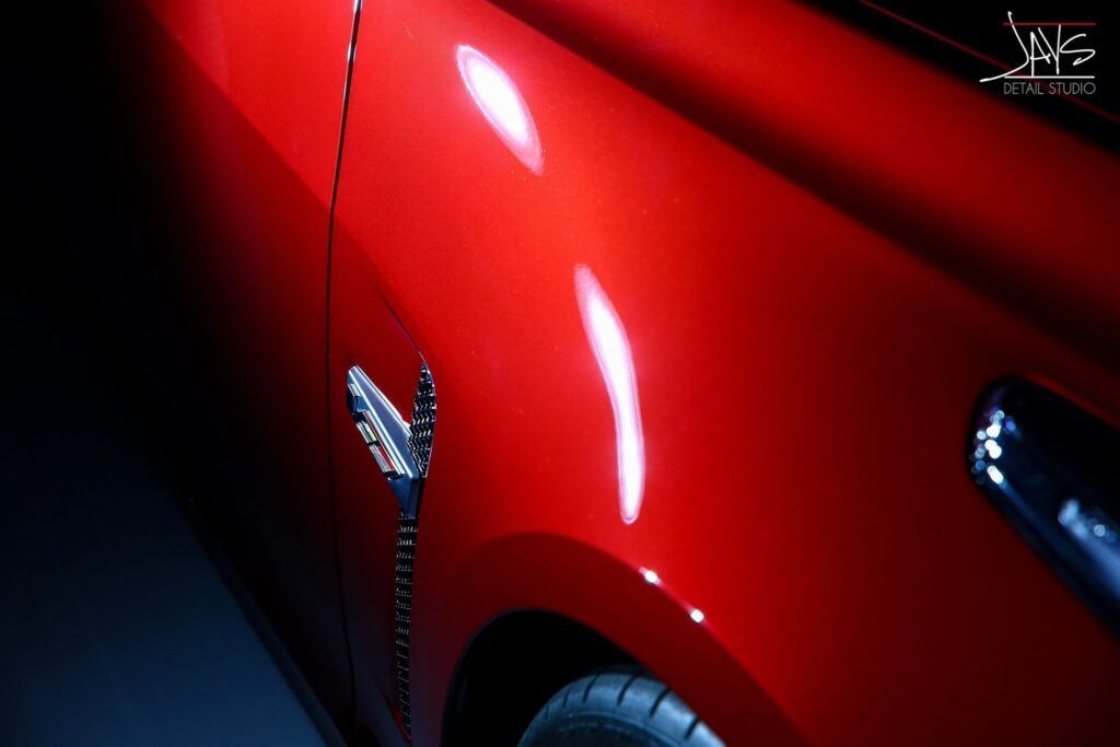 Red car's side, glossy finish, with a silver emblem and wheel detail visible. Lit dramatically against a dark background.