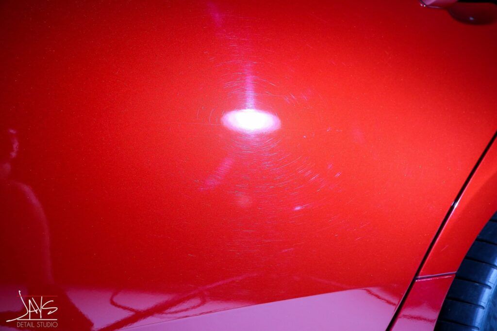 Red car paint with swirl marks, a bright reflection, and a tire visible.