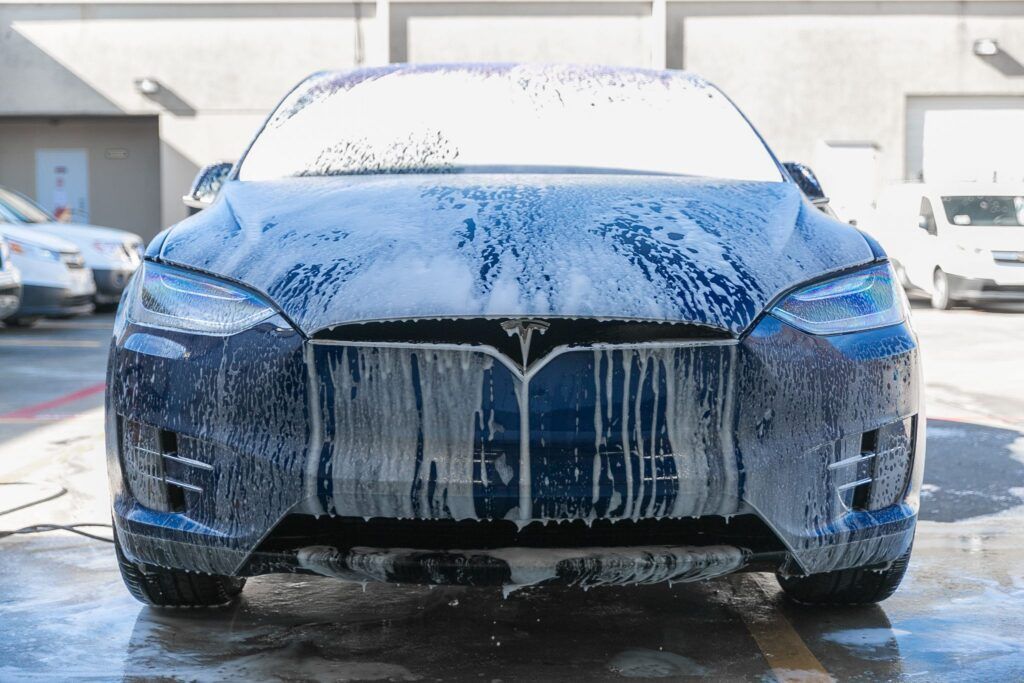 Blue Tesla car covered in white soap suds at a car wash.