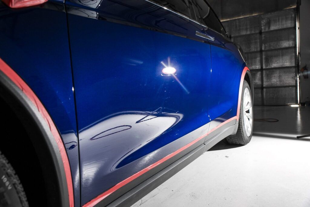 Blue car's side with red protective tape, in a garage. Shiny paint reflects light.