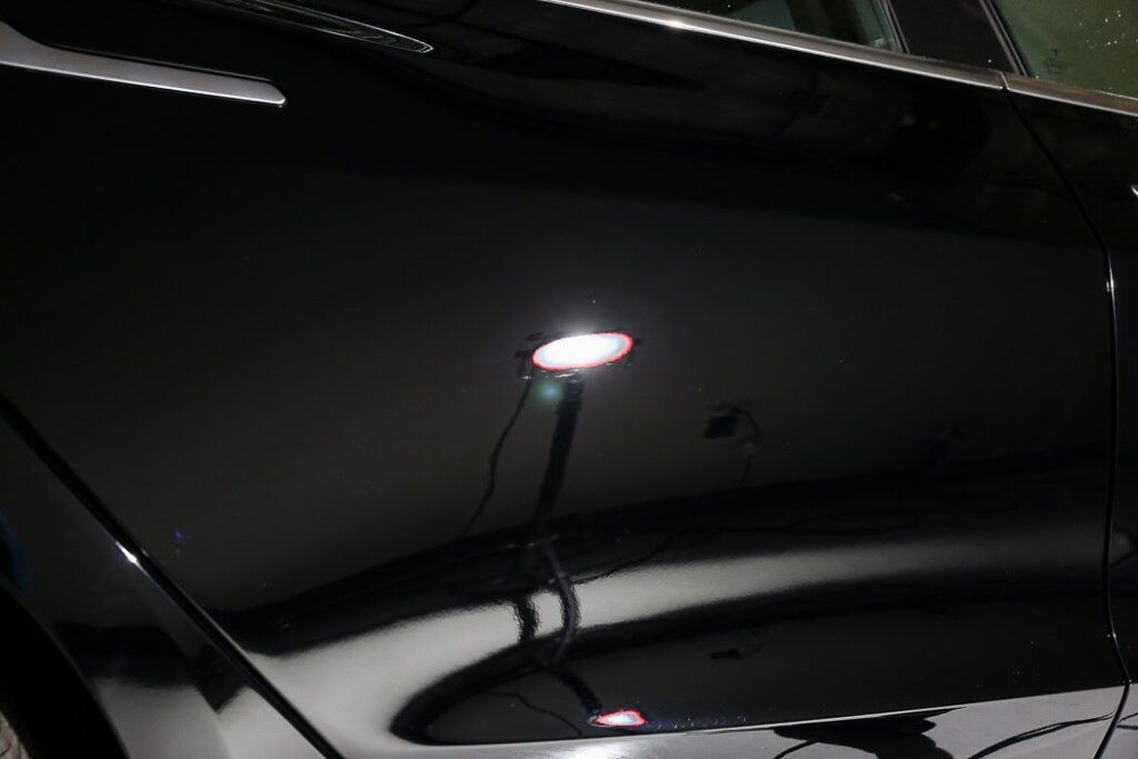 Black car door reflecting a bright lamp, showing surface imperfections in the paint.