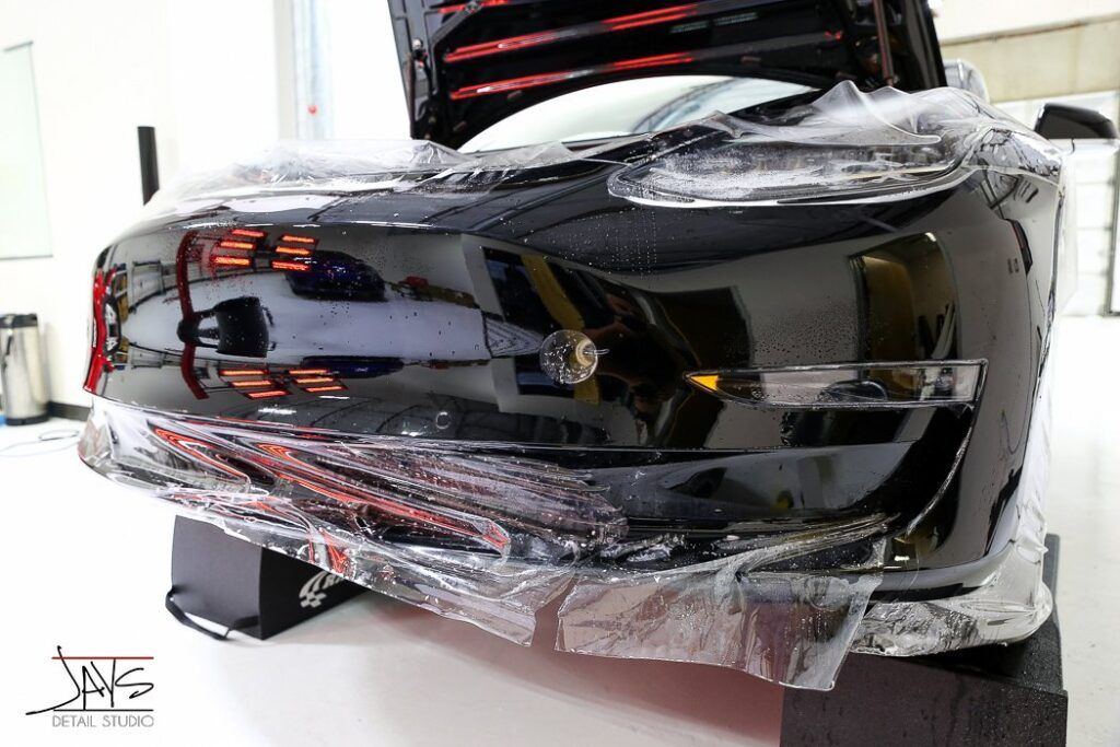 Black car with protective film on its front, inside a brightly lit garage.