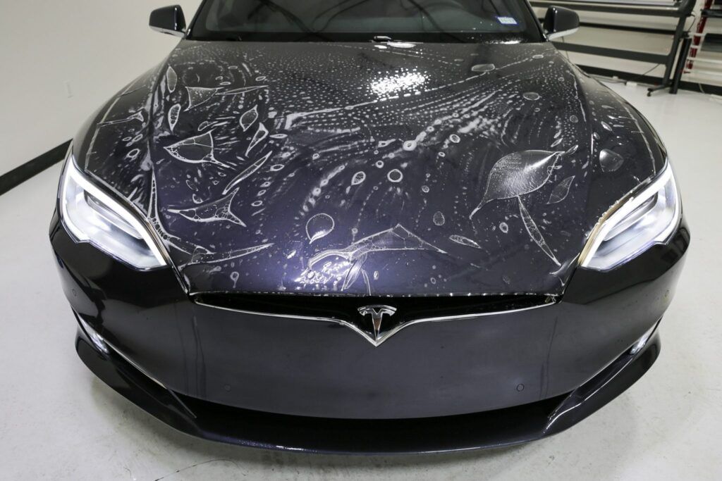 A dark Tesla with wet, protective film on the hood.