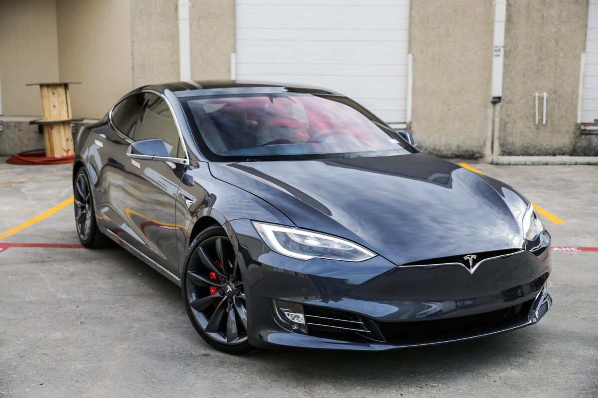 Dark gray Tesla Model S parked in front of a building with its headlights on.