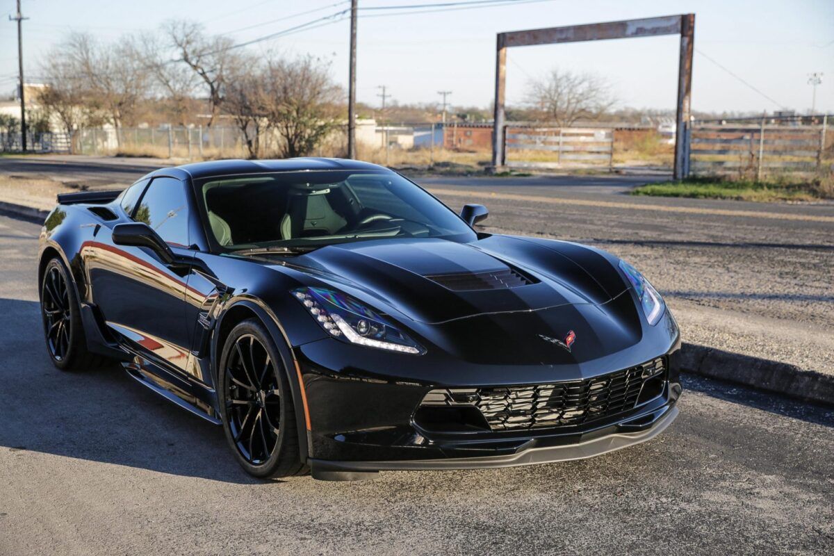 Black Chevrolet Corvette sports car parked on a paved street in front of a fence.