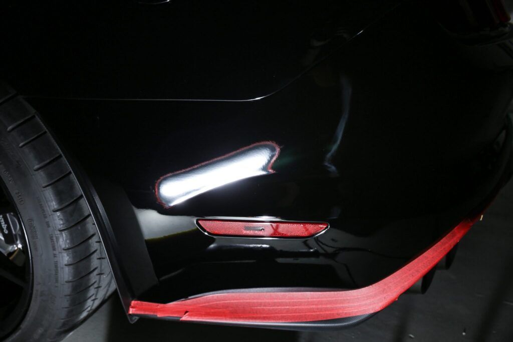 Black car's rear bumper with a red reflector and accent, part of the tire and wheel visible.