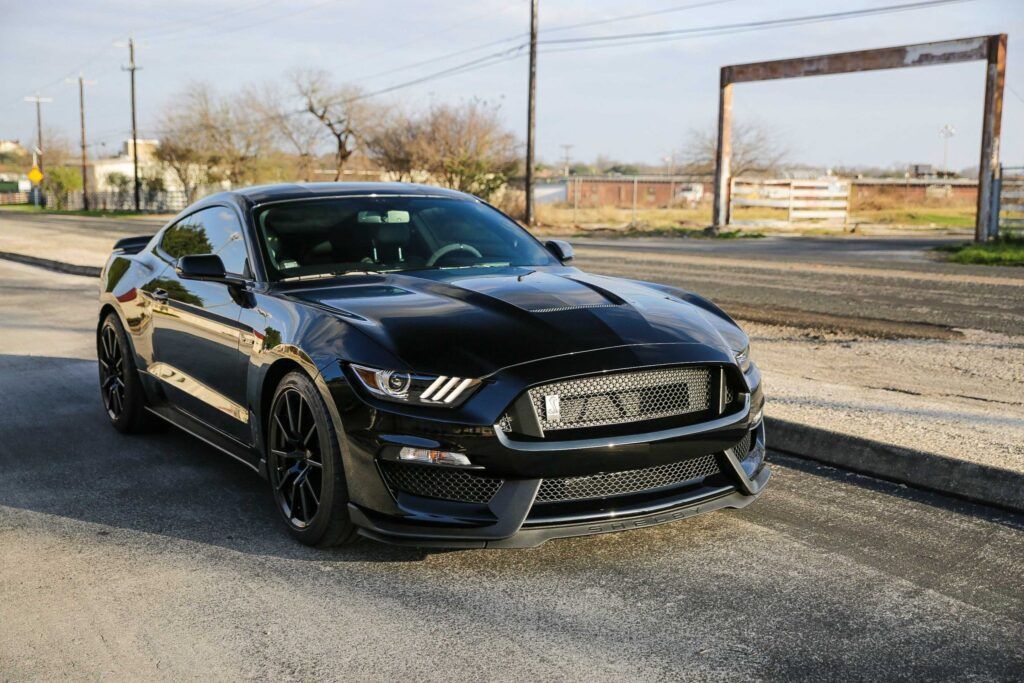 Black Ford Mustang sports car parked on a road, near a fence, sunny day.