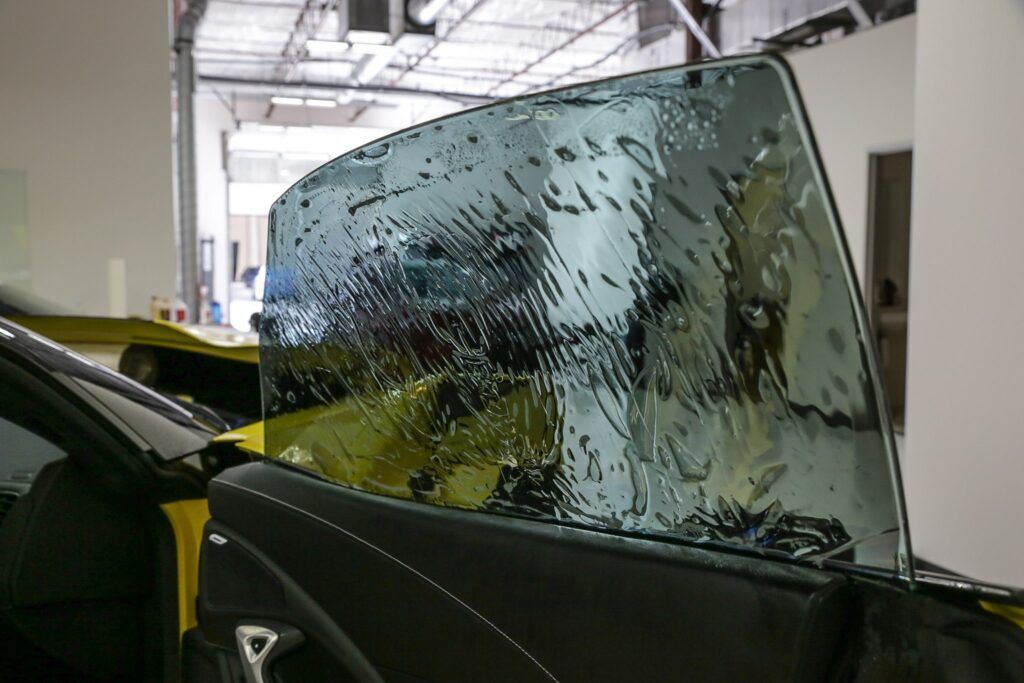Car window with wet tint film being applied, yellow car in a shop.