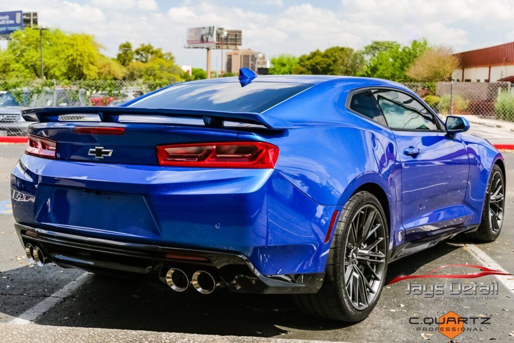 Blue Chevrolet Camaro sports car, rear view, parked. Black rims, dual exhaust, spoiler, in a parking lot.