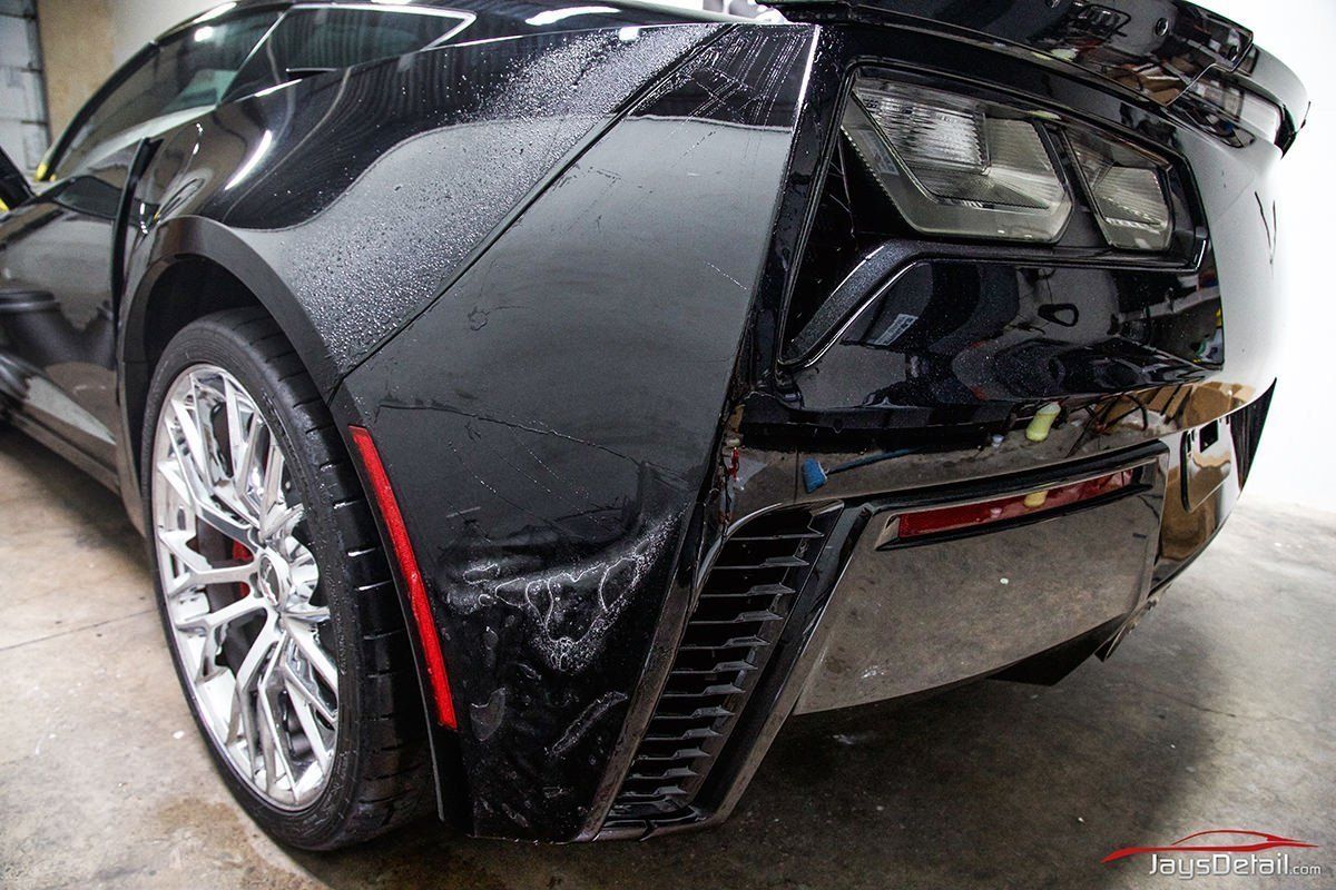 Black sports car's rear, wet surface, chrome wheels, red details, indoor setting.