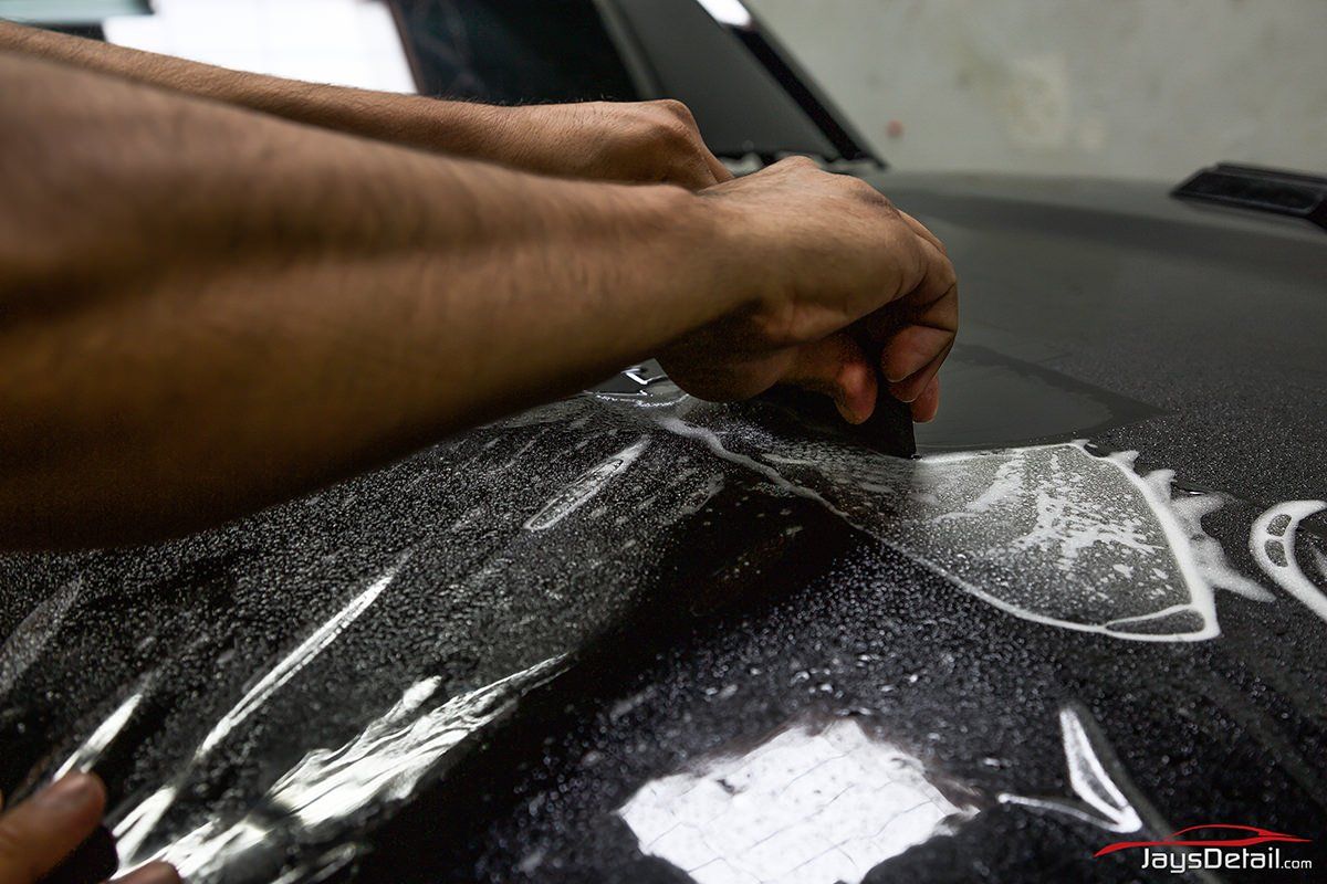 Hands applying protective film to a black car hood in a garage setting.