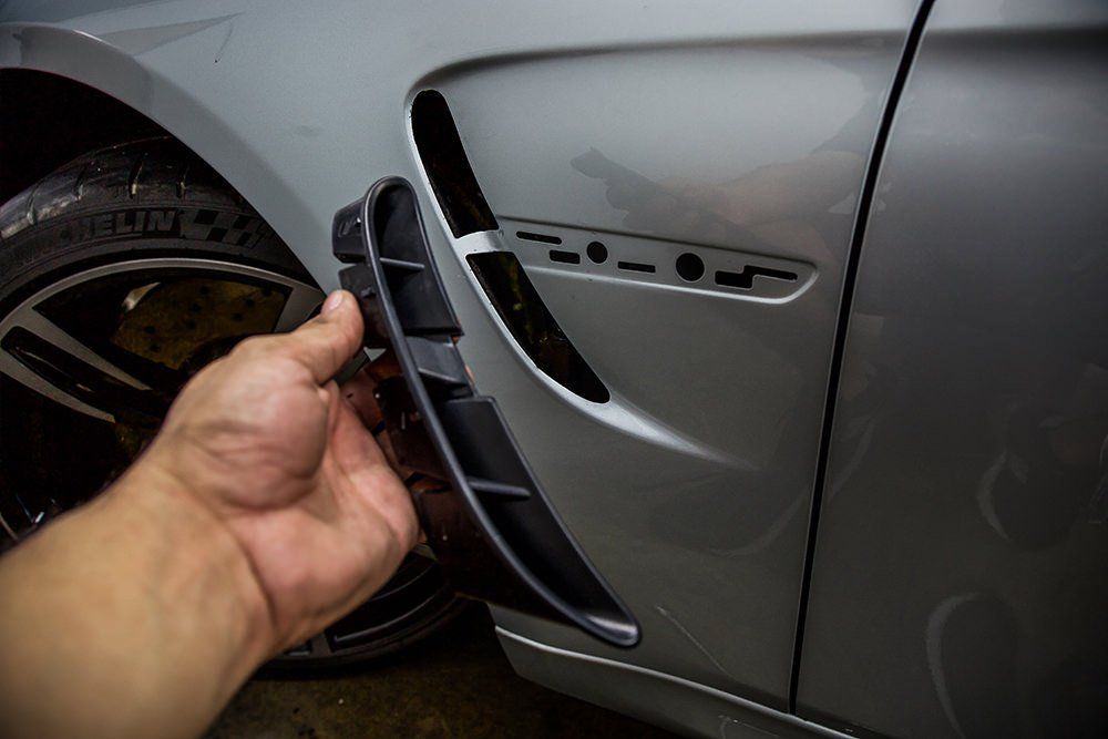 A person holding a black vent cover next to a silver car's front fender, showing removal of the part.