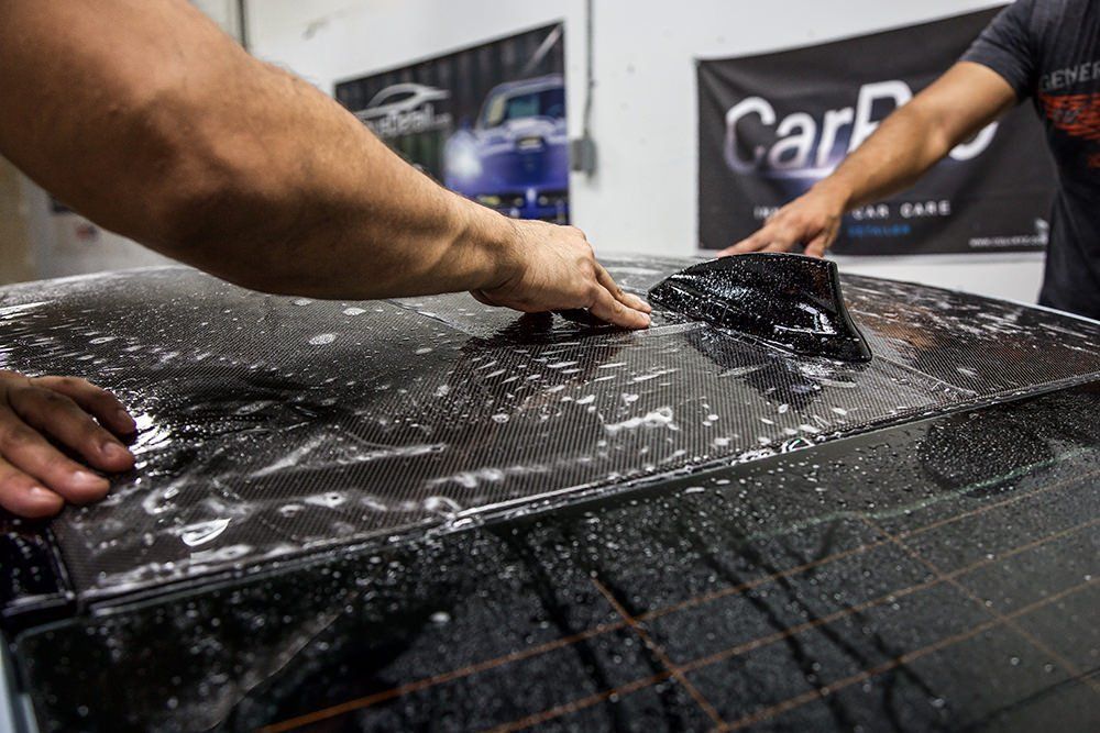 Two people applying film to a car's black roof, soapy water visible. Workshop setting with car care banners.