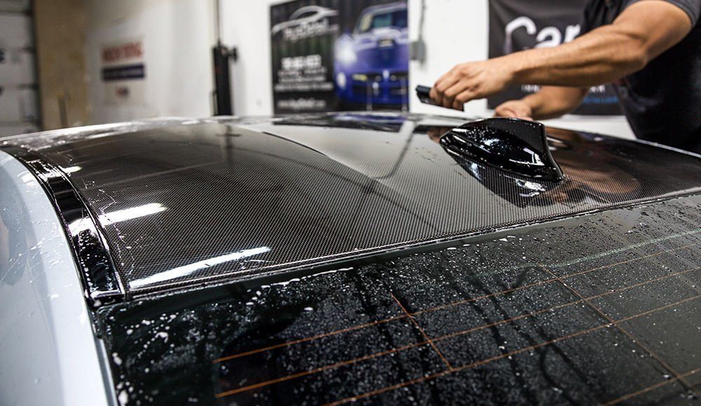 Person applying tint to a car's black roof. Water sprayed, tint applied; indoor shop setting.