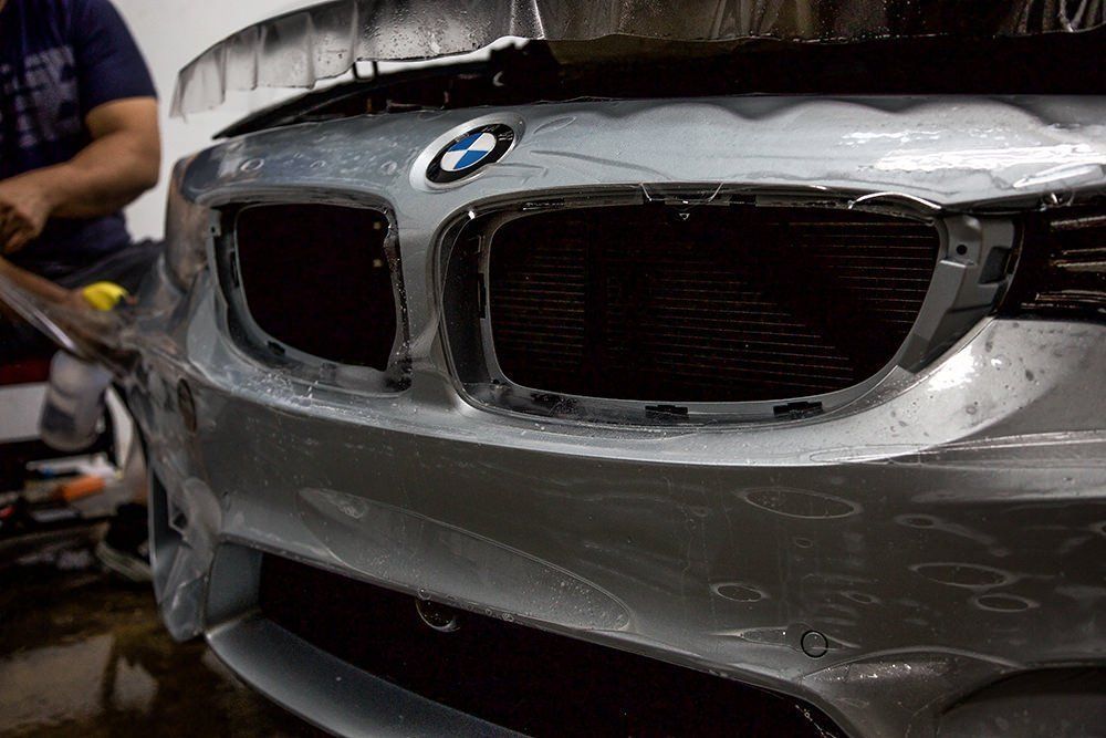 Silver BMW bumper being wrapped with protective film. Man in background.
