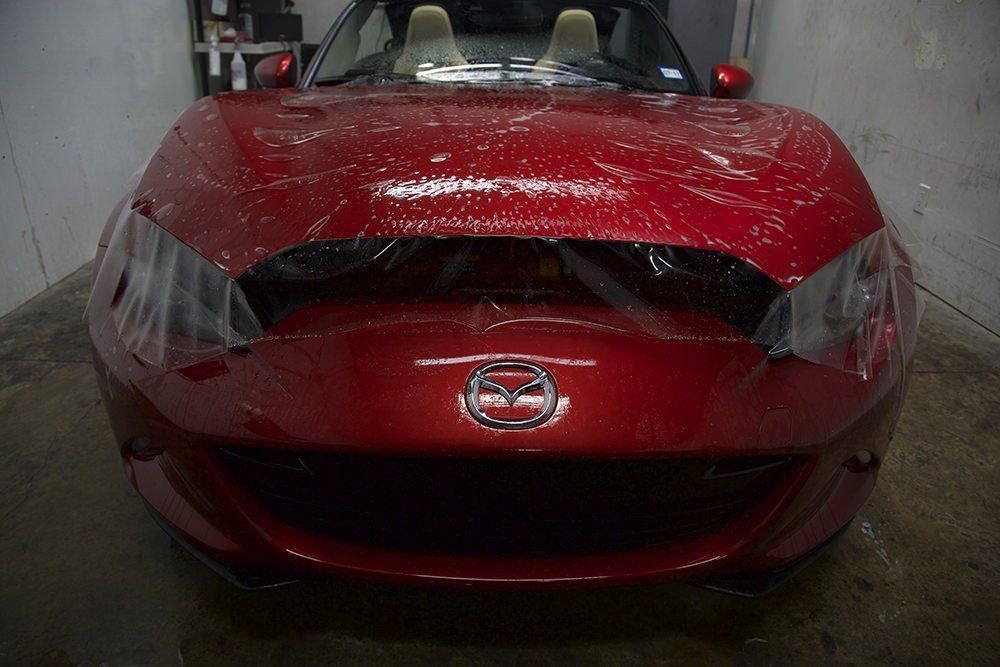 Red Mazda Miata with protective film on the hood, inside a garage.
