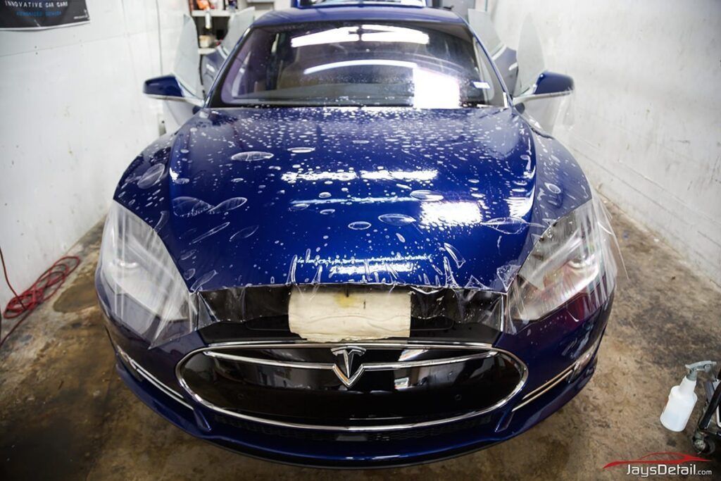 Blue Tesla car being worked on, with protective film being applied in a garage setting.