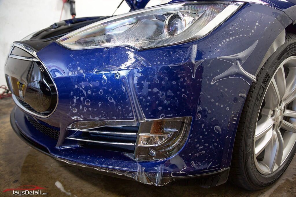 Blue Tesla car bumper with protective film being applied, water droplets visible.