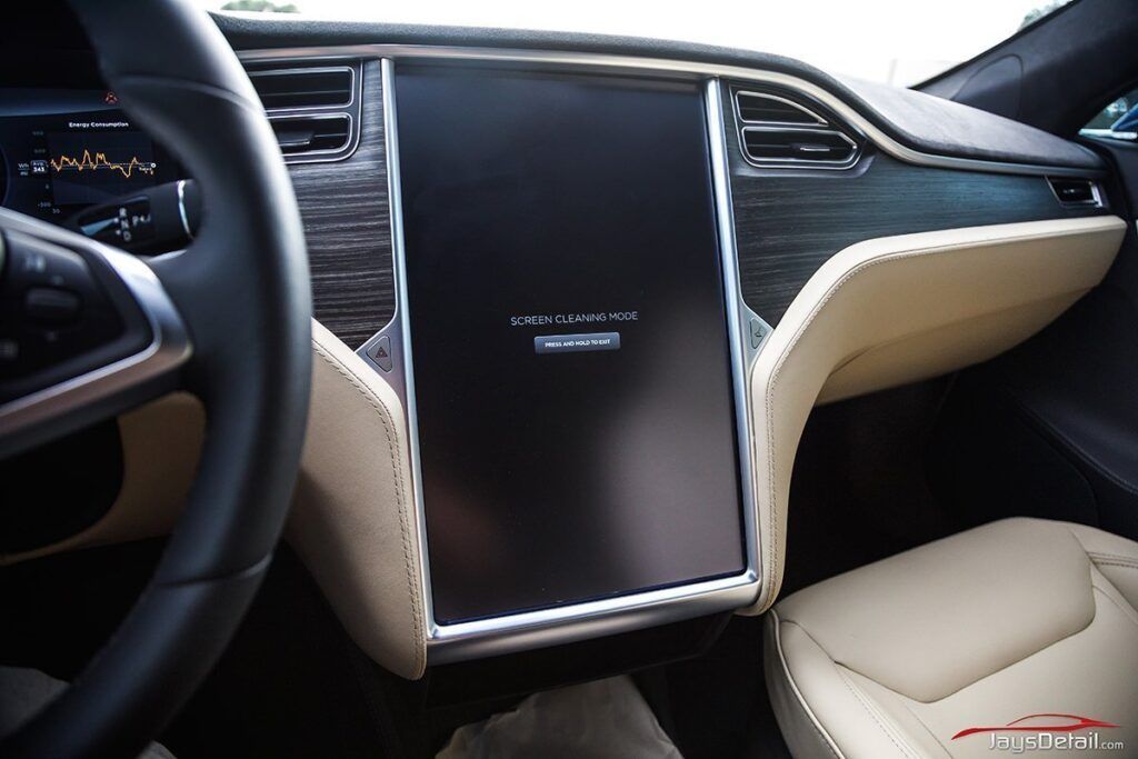 Interior of a Tesla car, center console with large touchscreen, wood trim, and cream-colored seats.