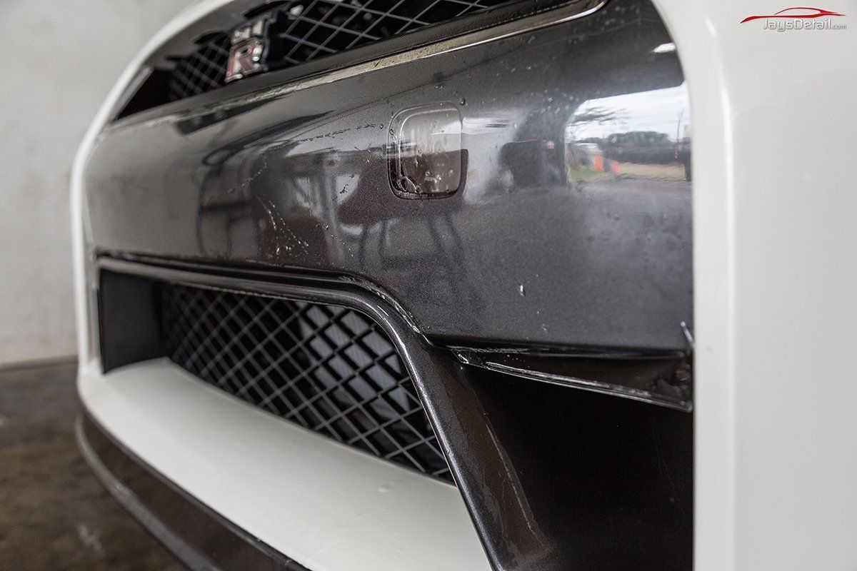 Close-up of a white car bumper with a carbon fiber grille, showing a damaged area.