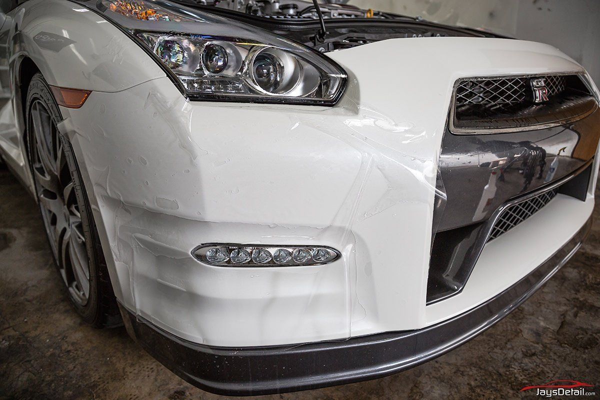 White Nissan GT-R sports car with protective wrap on the front bumper and hood, in a garage setting.
