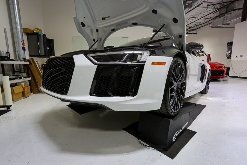 White and black Audi R8 with open hood on black ramps in a shop.