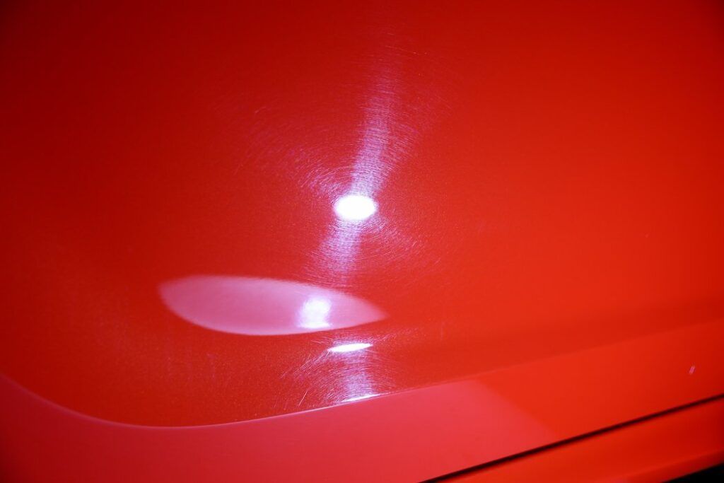 Close-up of a shiny red car surface, reflecting bright light with visible swirl marks.
