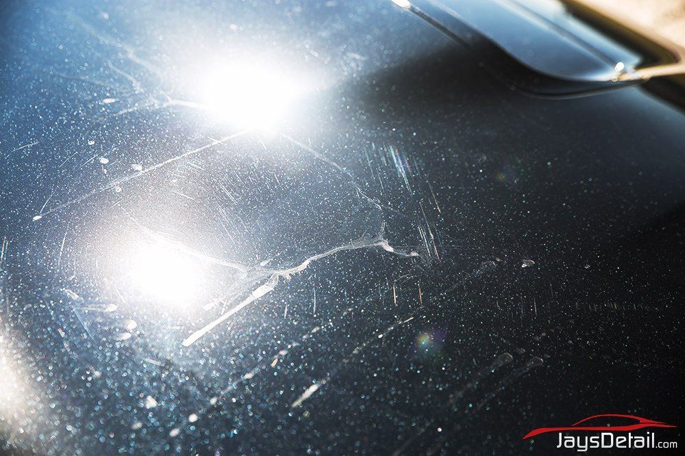 Scratched dark car hood with light reflections, showing damage and a cracked area.