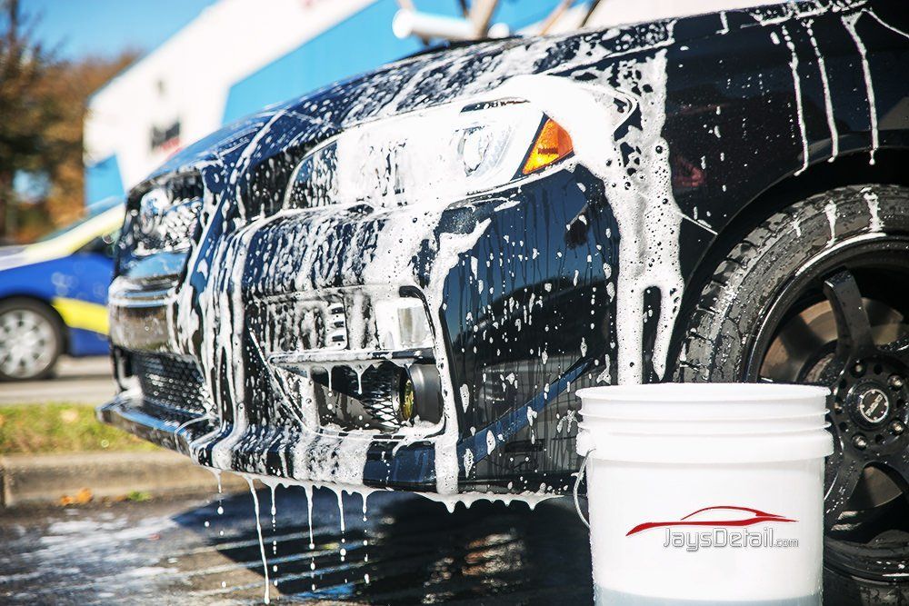 Black car covered in white soap suds at a car wash, with a bucket of soap and a tire visible.