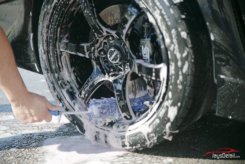 Person washing a black car's wheel with a brush and soapy water outdoors.